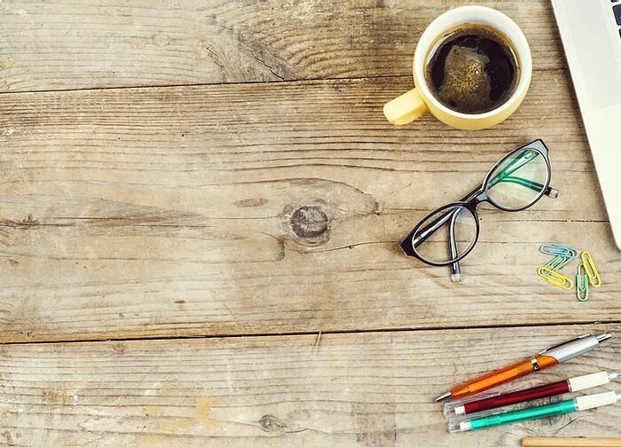 Laptop, cell phone, coffee cup, glasses, pens and paperclips sitting on table.